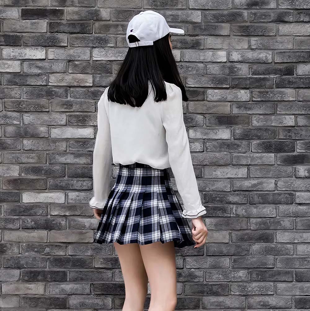 Femme vue de dos portant une jupe écossaise bleue et blanche à plis, style kilt, avec un haut blanc et une casquette blanche, devant un mur en briques grises