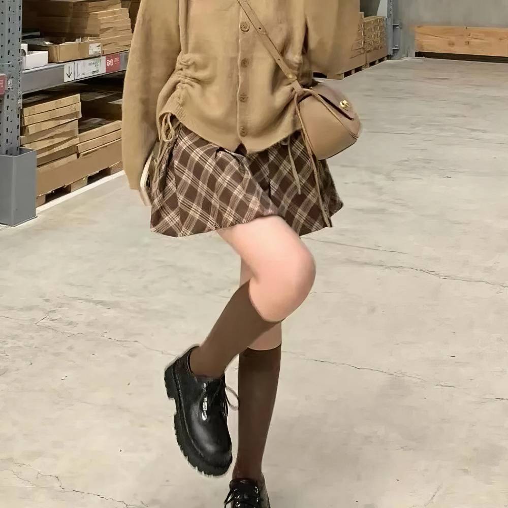 Mini jupe tartan écossaise plissée marron et beige portée avec pull marron et bottines noires dans un entrepôt